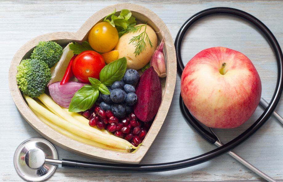 A Photograph of Fruits and Vegetables in a Heart Shaped Bowl. Wrapped Around the Bowl is a Stethoscope and an Apple